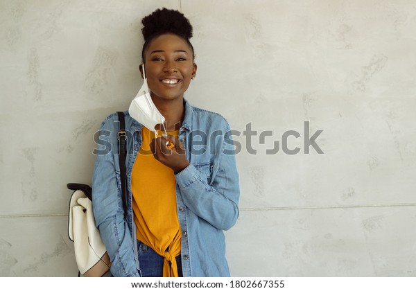 Happy black\
college student taking her mask off while standing against the wall\
and looking at camera. Copy\
space.