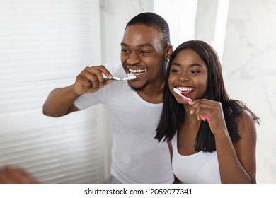 Happy Black Couple Brushing Teeth Near Mirror In Bathroom, Cheerful Young African American Spouses Making Morning Oral Hygiene Together, Holding Toothbrushes And Smiling To Their Reflection, Closeup - Powered by Shutterstock