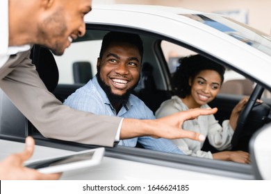 Happy Black Couple Buying New Car Testing And Talking With Salesman Sitting In Auto Dealership Showroom. Family Choosing Automobile.