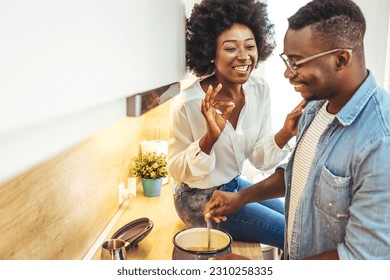 Happy black couple cooking and tasting healthy food in kitchen. Beautiful young couple is talking and smiling while cooking in kitchen at home. Man and woman in love cooking food together at home. - Shutterstock ID 2310258335