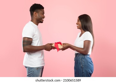 Happy Black Couple Giving Gifts To Each Other, Pink Studio Background, Free Space. Loving Handsome Man Giving Present To His Excited Wife Or Girlfriend. Valentines Day Celebration, Profile Side View