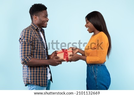 Happy black couple giving gifts to each other, blue studio background, free space. Loving handsome man giving present to his excited wife or girlfriend. Valentines day celebration, profile side view