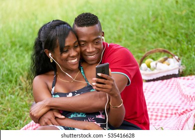 Happy black couple having fun in city park. Young african american man and woman listening to music and watching video on mobile phone.