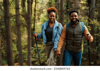 Happy black couple hiking in a lush green forest, holding hands and smiling. They are dressed in casual outdoor clothing, with hiking poles in hand. - Powered by Shutterstock