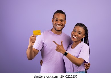 Happy Black Couple Holding And Pointing Finger At Credit Card Recommending Service Smiling To Camera Over Purple Background. Excited Bank Customers Showing Card For Payments Posing In Studio