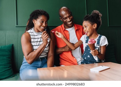 A happy black couple laughs with their daughter, who is eating an ice cream cone. They are sitting in a cozy cafe, sharing ice cream together. - Powered by Shutterstock