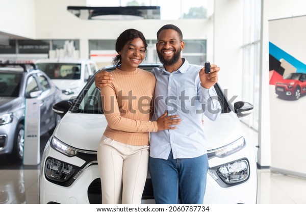 Happy Black Couple Posing With Car Keys In Hands In\
Modern Dealership Center, Cheerful African American Spouses\
Standing Near Their New Vehicle In Showroom And Smiling At Camera,\
Copy Space