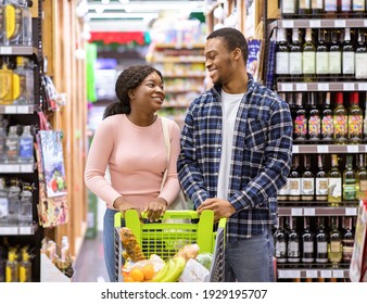 Happy Black Couple With Shopping Cart Looking At Each Other And Smiling At Alcohol Department Of Supermarket. Positive Young Family Buying Beverages At Liquor Store Or Mall