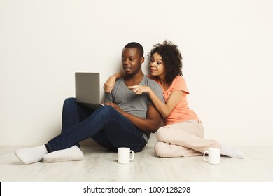 Happy Black Couple Shopping Online, Making Order With Laptop While Sitting At Home Floor In Empty Living Room, Copy Space, Isolated