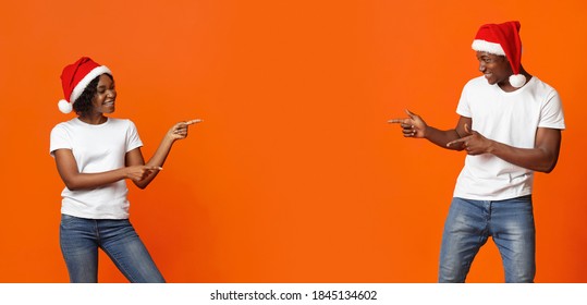 Happy Black Couple Showing Advert For New Year Or Christmas Deal, Offer, Sale, Discount. Emotional African American Young Man And Woman In Santa Hats Pointing At Copy Space On Orange Studio Background