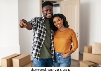 Happy Black Couple Showing New House Key Posing Embracing Standing Among Cardboard Moving Boxes At Home. Selective Focus On Keys. Real Estate Ownership, Family Housing