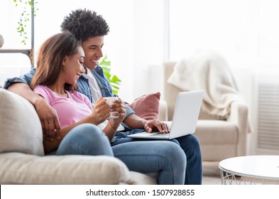 Happy black couple sitting on couch watching movie on laptop, drinking coffee and relaxing at home, side view with copy space - Shutterstock ID 1507928855