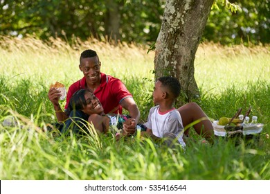 Happy Black Couple With Son In City Park. African American Family With Young Man, Woman And Child Eating Food During Picnic.