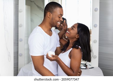 Happy Black Couple Spending Time In Bathroom Together, Caring Wife Applying Cream On Husband's Face And Laughing, Romantic Young African American Spouses Making Beauty Routine At Home, Free Space - Powered by Shutterstock