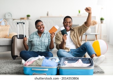 Happy Black Couple Of Travelers Gesturing Yes Posing With Travel Tickets Sitting With Packed Suitcase Ready For Summer Trip At Home, Smiling To Camera. Tourists Celebrating Long-Awaited Vacation