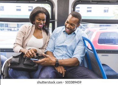 Happy Black Couple Travelling By Bus In Chicago. A Man And A Woman Sitting In The Bus And Looking At The Smartphone. They Are Laughing And Enjoying Time Together. Lifestyle And Happiness Concepts.