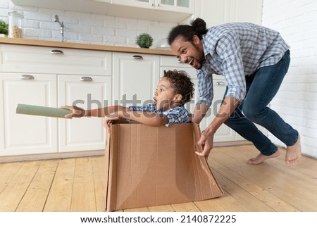 Happy Black dad driving cardboard box with excited little son inside, playing with kid at home, enjoying active games, leisure, entertainment after moving into new apartment. Family, relocation