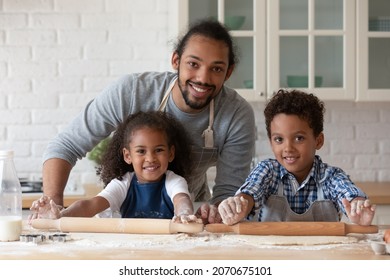 Happy Black Dad Helping Children In Aprons To Bake Pie, Cake, Rolling Dough On Flour Table, Looking At Camera, Smiling. Little Kids And Dad Cooking Pastry For Family Dinner. Head Shot Portrait