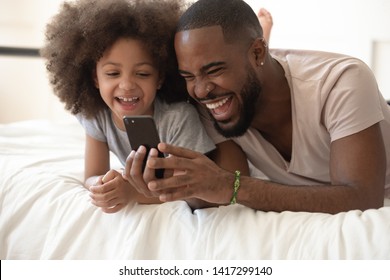 Happy Black Dad Use Funny App Laugh Lying On Bed With Cute Little Kid Daughter, Cheerful African Family Father And Small Child Girl Holding Phone Looking At Screen Having Fun In Bedroom In Morning