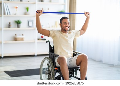 Happy Black Disabled Man In Wheelchair Doing Exercises With Rubber Band At Home. African American Handicapped Guy Working Out Indoors, Keeping In Grey Shape, Enjoying His Training