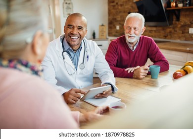 Happy black doctor using touchpad and communicating with a senior couple while visiting them at home.