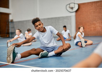 Happy Black Elementary Student Stretching His Leg While Warming Up During Physical Education Class At School Gym And Looking At Camera. 