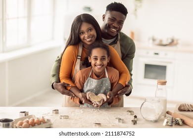 Happy Black Family Baking Showing Heart-Shaped Dough In Hands Smiling To Camera Standing In Kitchen At Home. Parents And Daughter Cooking Making Pastry. Food Recipes Concept