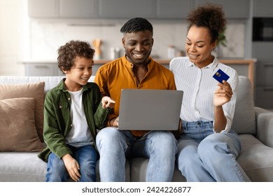 Happy black family with boy enjoying online shopping, sitting on couch with laptop as the mother holding credit card in bright living room interior - Powered by Shutterstock