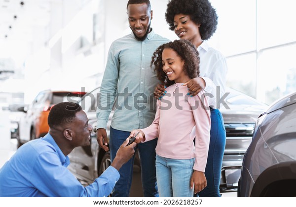 Happy black family\
buying new car, salesman giving them key at auto dealership. Afro\
clients making deal with manager, purchasing modern vehicle at\
automobile showroom