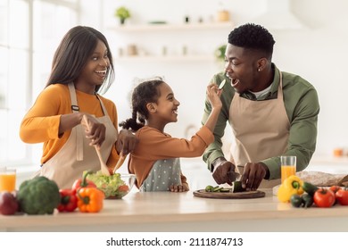 Happy Black Family Cooking Having Fun, Daughter Feeding Father While Making Vegetable Salad For Dinner Standing In Modern Kitchen Indoors. Joyful Parents And Daughter Preparing Healthy Food