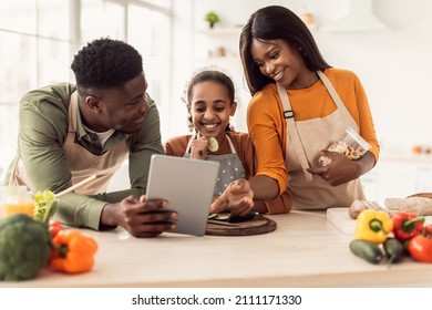 Happy Black Family Cooking Using Digital Tablet Computer Preparing Dinner Together Browsing Recipes Online In Modern Kitchen At Home. Parents And Daughter Making Salad