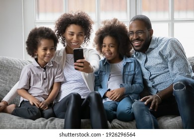 Happy Black Family Couple And Two Kids Taking Selfie Picture On Smartphone At Home. Millennial African American Parents And Children Using Cell Phone For Video Call And Virtual Chat With Grandparents