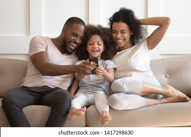 Happy Black Family Having Fun Play Mobile Game, Take Selfie, Watch Funny Video On Phone, Cute Little African Kid Daughter And Parents Holding Looking At Smartphone Laugh Enjoy Using App Sit On Sofa