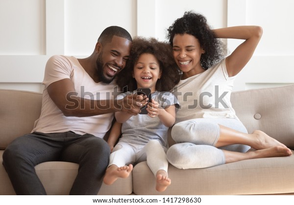 楽しいゲームを楽しむ黒人家族 自撮り 携帯電話で面白いビデオを見る かわいい小さなアフリカの子どもの娘 スマートフォンを見ている両親 ソファーのアプリの座で楽しむ の写真素材 今すぐ編集