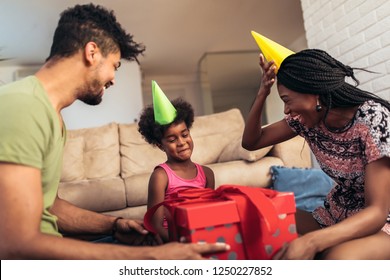 Happy Black Family At Home. African American Father, Mother And Child Celebrating Birthday, Having Fun At Party. Young Woman Giving Gift To Daughter