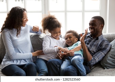 Happy Black Family Laughing Playing With Children At Home, Cheerful African American Parents Tickling 2 Mixed Race Kids Having Fun On Couch, Mom Dad With Little Son Daughter Enjoying Funny Activity