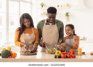 Happy Black Family Making Salad Adding Oil, Mixing Ingredients In Bowl And Cutting Vegetables In Modern Kitchen Indoor. Daughter Helping Parents Prepare Dinner At Home. Healthy Nutrition