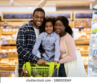 Happy black family with shopping cart purchasing food at supermarket. Cheerful African American parents with their daughter posing and looking at camera in huge mall