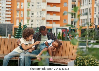 Happy Black Family Sitting And Resting On Wooden Bench In Yard Of Modern District Outdoors. Father Hugging His Laughing Daughter And Son. Fatherhood And Parenting. Relationship And Enjoy Time Together