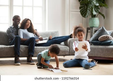 Happy black family spend free time together in living room at home. Couple sitting on sofa looks at little daughter show her drawing, son have a fun on warm wooden floor drawing with colorful pencils