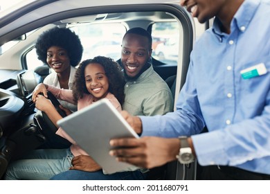 Happy Black Family Testing New Car, Salesman Showing Them Purchase Or Lease Agreement On Digital Tablet At Auto Dealership. Afro Clients Making Deal With Manager, Buying Vehicle At Showroom