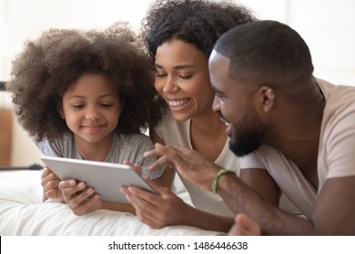 Happy black family of three lying on bed, using digital tablet together. Smiling african american girl with loving parents watching cartoons, photos or shopping, making order, ecommerce concept. - Powered by Shutterstock