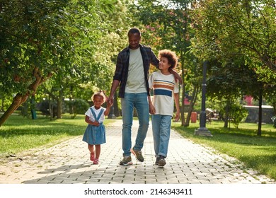 Happy Black Family Walking On Pavement Road In Green Park. Father Hugging Son And Holding Hand Of Little Daughter. Relationship And Enjoying Time Together. Fatherhood And Parenting. Sunny Summer Day