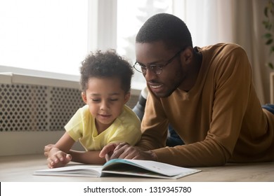 Happy Black Father Baby Sitter And Toddler Son Having Fun With Book Lying On Warm Floor, African Dad Teaching Little Kid Boy Learning To Read, Parent With Children Educational Activities At Home