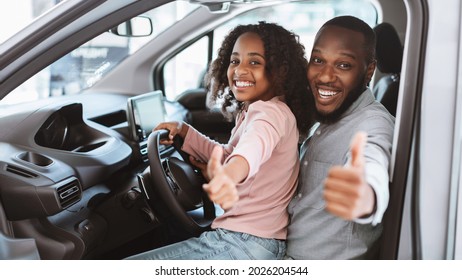 Happy Black Father And His Daughter Sitting On Driver Seat Of New Car, Showing Thumb Up Gesture, Recommending Auto Dealership. Cheerful Afro Family Test Driving Vehicle Together At Automobile Showroom