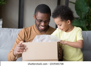 Happy black father and little kid son opening cardboard box looking inside, african american dad with cute child toddler clients receiving carton package, post mail parcel delivery service concept