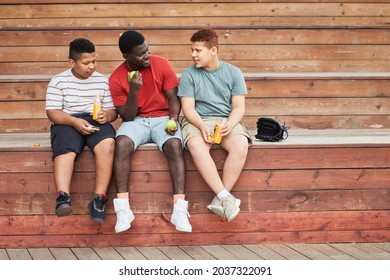 Happy Black Father Sitting On Bleacher Bench And Holding Tennis Balls While Chatting With Sons Drinking Juice