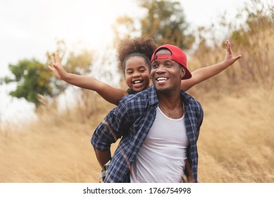 Happy Black Father Smiling Having Fun And Giving Piggyback Ride To Joyful Daughter In Autumn At Park On Holiday. African Man And African-American Girl Enjoying In Dry Field Nature. Family Lifestyle