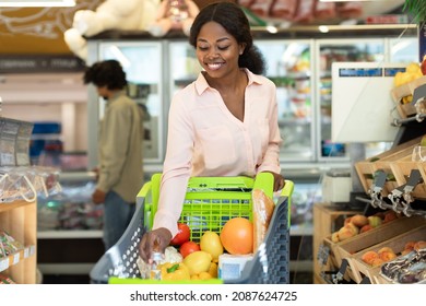 Happy Black Female Buyer Walking With Shop Cart Full Of Food Doing Grocery Shopping In Modern Supermarket On Weekend. Lady Buying Products In Groceries Store Concept
