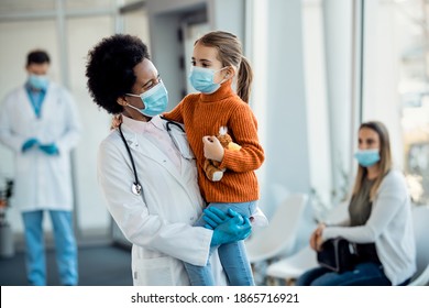 Happy Black Female Doctor Holding Little Girl While Standing In A Lobby At The Hospital. Both Of Them Are Wearing Protective Face Masks. 